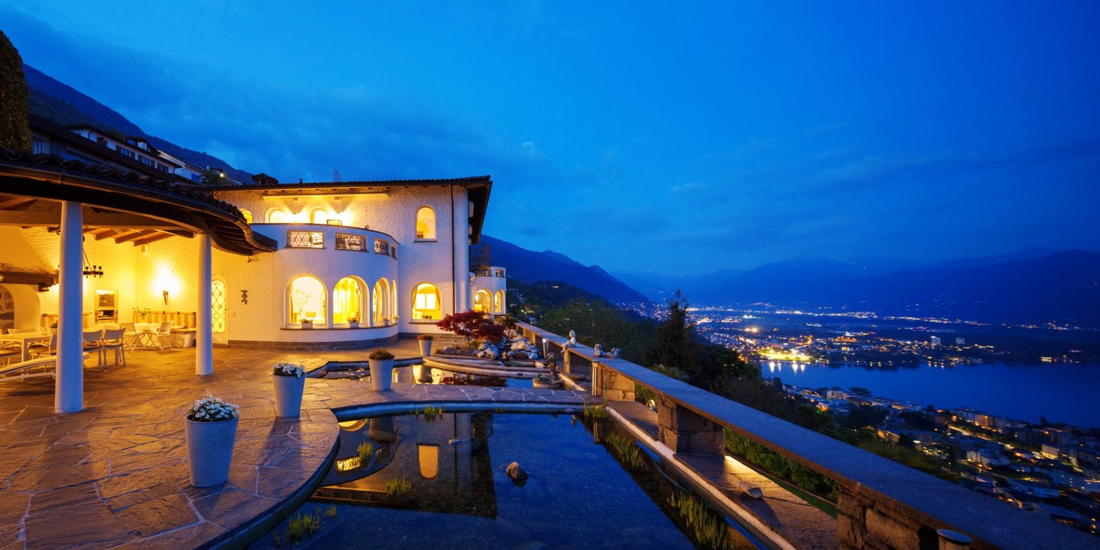 The terrace of a villa at dusk, above a lake and the lights of the town